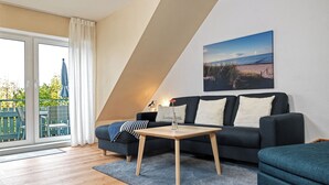 A modern living room with a dark sofa, a wooden coffee table, and a view of the outdoors through a large window. - Apartment Haffblick - Beautiful holiday apartment for up to 4 people. (Insel Poel / OT Kaltenhof)