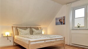 A bedroom with a wooden bed, white walls, a window with blinds, and a small potted plant on the windowsill. - Apartment Haffblick - Beautiful holiday apartment for up to 4 people. (Insel Poel / OT Kaltenhof)