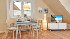 A dining area with a table set for four, a hanging light fixture, a television on a white cabinet, and framed pictures on the wall. - Apartment Haffblick - Beautiful holiday apartment for up to 4 people. (Insel Poel / OT Kaltenhof)
