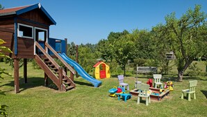 A wooden playhouse with a slide, a small red and yellow playhouse, and a picnic table with chairs and toys. - Apt. WINDROSE - Cozy semi-detached house for 4 people (Insel Poel / OT Gollwitz)