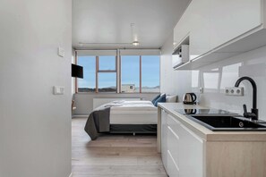 A modern bedroom with a bed, bedside table, and a view of the outdoors through large windows. - Iceland Comfort Apartments Economy Studio (Kópavogur)