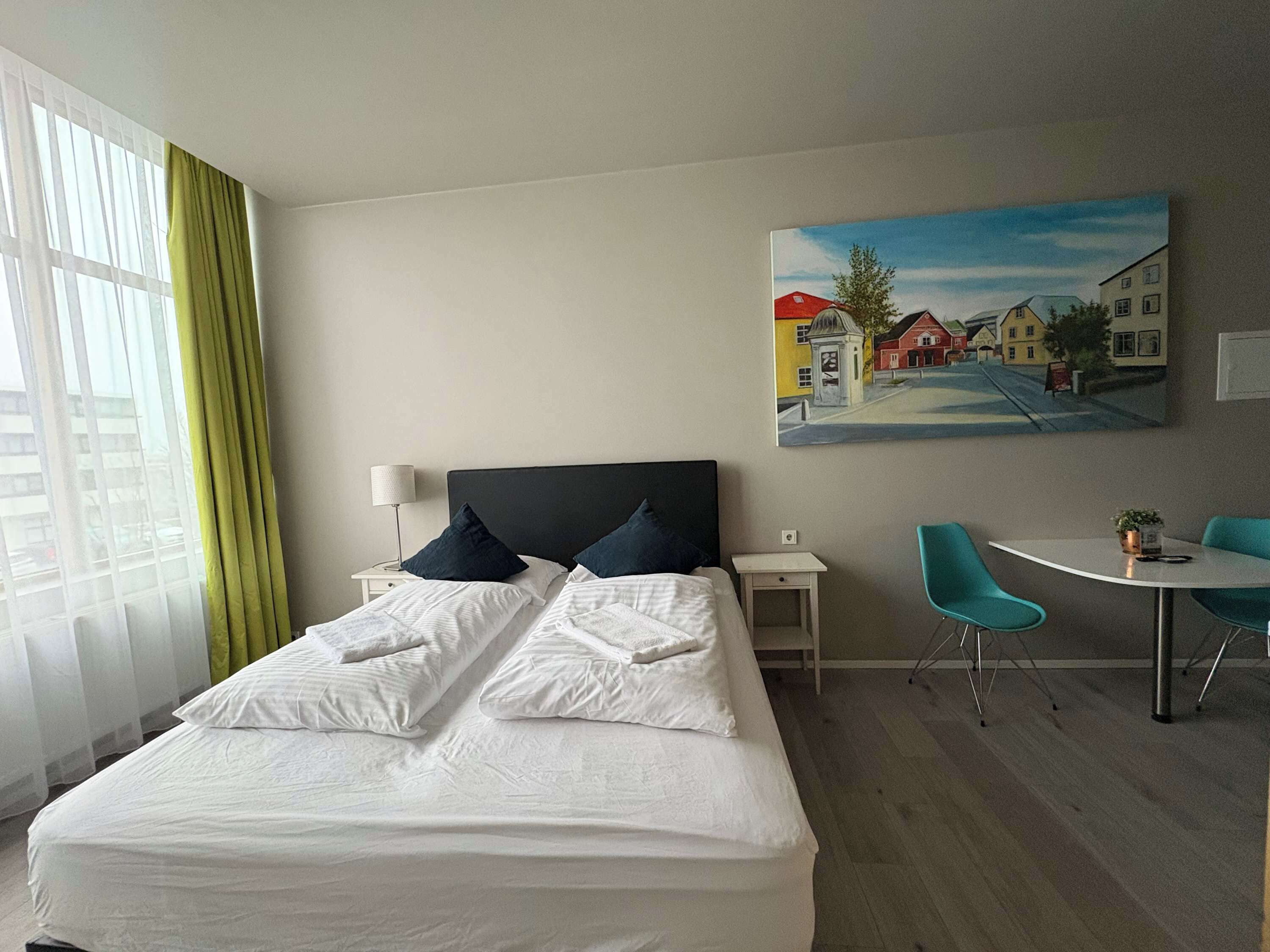 Et hotelværelse med en seng, to sengeborde, et skrivebord med stole, et billede på væggen og et vindue med gardiner.