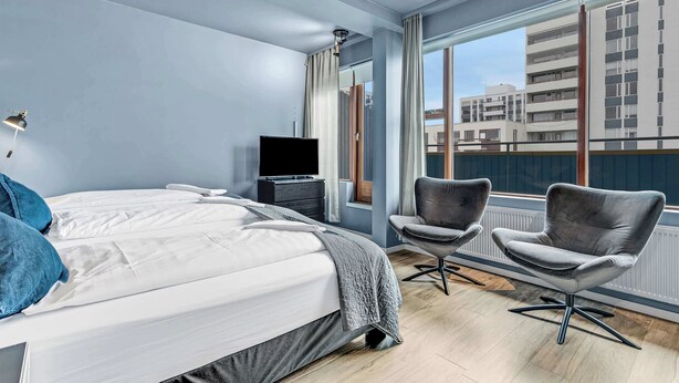 A modern hotel room with a large bed, two armchairs, a TV, and a view of buildings through large windows.