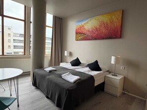 Ein Hotelzimmer mit Bett, Nachttischen, einem Schreibtisch und einem Fenster mit Blick auf Gebäude.