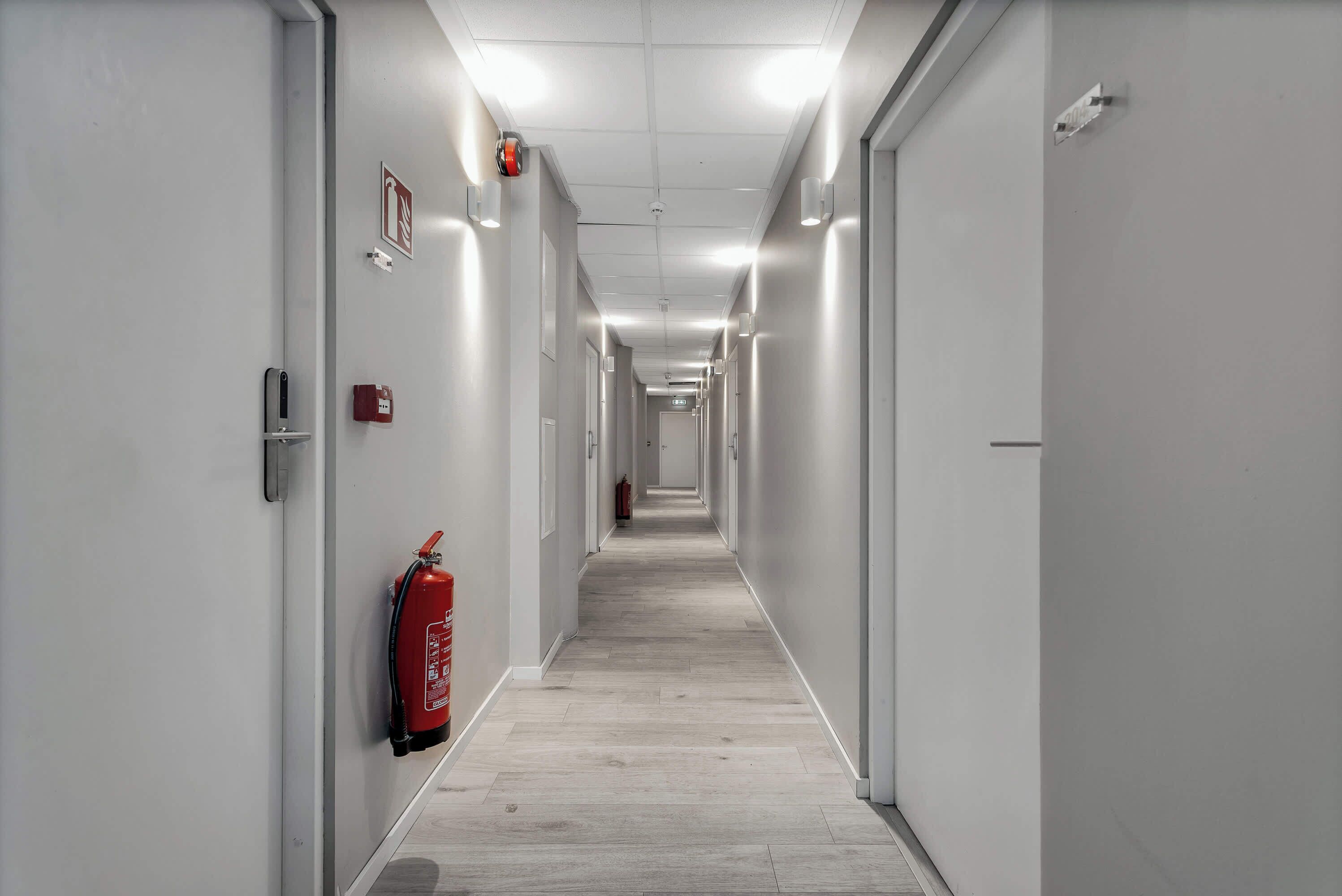 A long, narrow hallway with white walls, a fire extinguisher mounted on the left wall, and a door at the end.