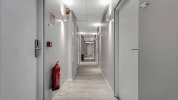 A long, narrow hallway with white walls, a fire extinguisher mounted on the left wall, and a door at the end.