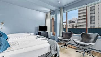A modern hotel room with a large bed, two armchairs, a TV, and a view of buildings through large windows.