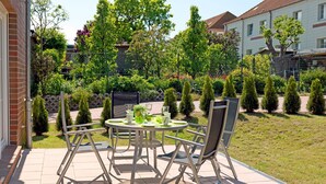 A patio with a round table and chairs, surrounded by a garden with various plants and a fence.