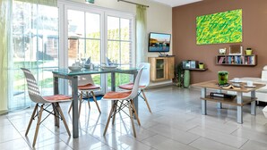 A modern living room with a dining area, large windows, a flat-screen TV, and a bookshelf.