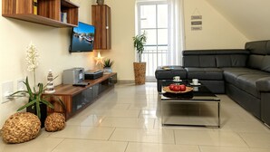 Een moderne woonkamer met een flatscreen televisie, houten planken, een zwart leren bankstel, een glazen salontafel met fruit en een groot raam met gordijnen.