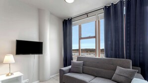 Una sala de estar moderna con un sofá gris, un televisor de pantalla plana, una mesita blanca y una ventana con cortinas.
