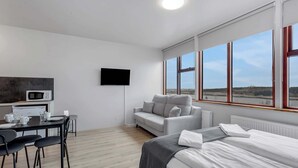 Una habitación de hotel moderna con zona de comedor, sofá, cama, televisor y ventanas grandes.