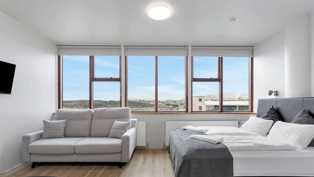 Un dormitorio moderno con cama, sofá, televisor y ventanas grandes con vista a la ciudad.