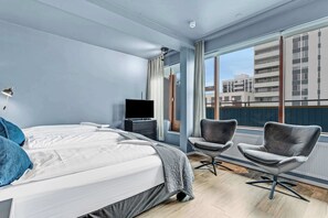 Une chambre d’hôtel moderne dotée d’un grand lit, de deux fauteuils, d’une télévision et offrant une vue sur les bâtiments grâce à de grandes fenêtres.
