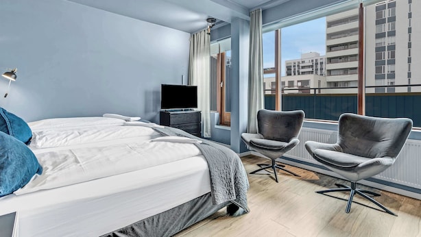 A modern hotel room with a large bed, two armchairs, a TV, and a view of buildings through large windows. - Iceland Comfort Apartments Triple Studio (Kópavogur)