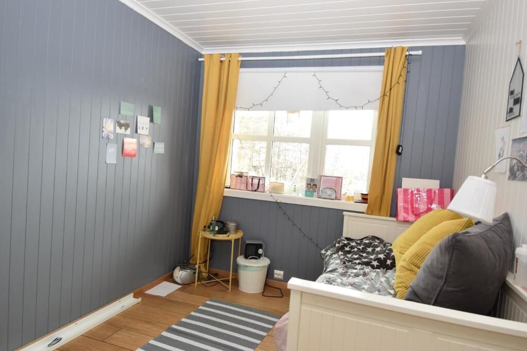 A small, cozy room with a bed, a window with curtains, a radiator, and a shelf with various items.