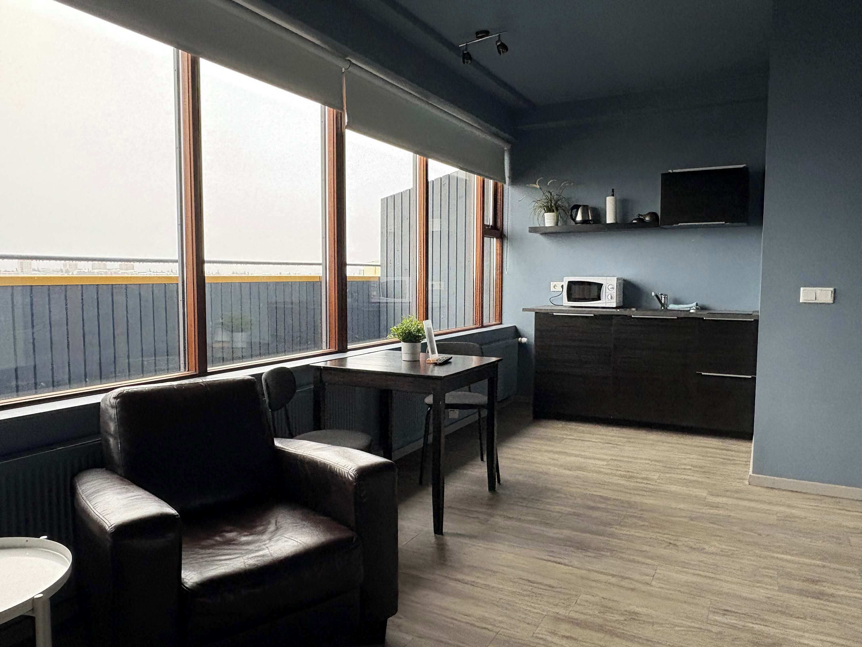 Une chambre moderne dotée d’une grande fenêtre, d’une table à manger, d’une chaise, d’un coin cuisine avec un four à micro-ondes et d’un téléviseur fixé au mur.