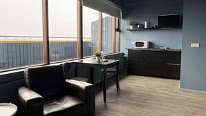 Una habitación moderna con un ventanal grande, una mesa de comedor, una silla, una zona de cocina con microondas y un televisor en la pared.