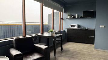 Une chambre moderne dotée d’une grande fenêtre, d’une table à manger, d’une chaise, d’un coin cuisine avec un four à micro-ondes et d’un téléviseur fixé au mur.