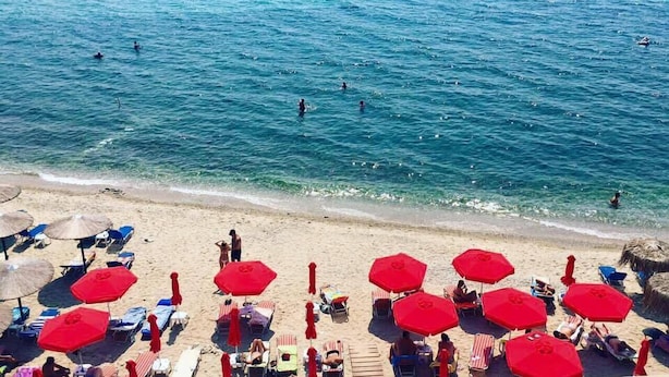 Una platja amb aigua blava cristal·lina, persones nedant i para-sols vermells fent ombra.