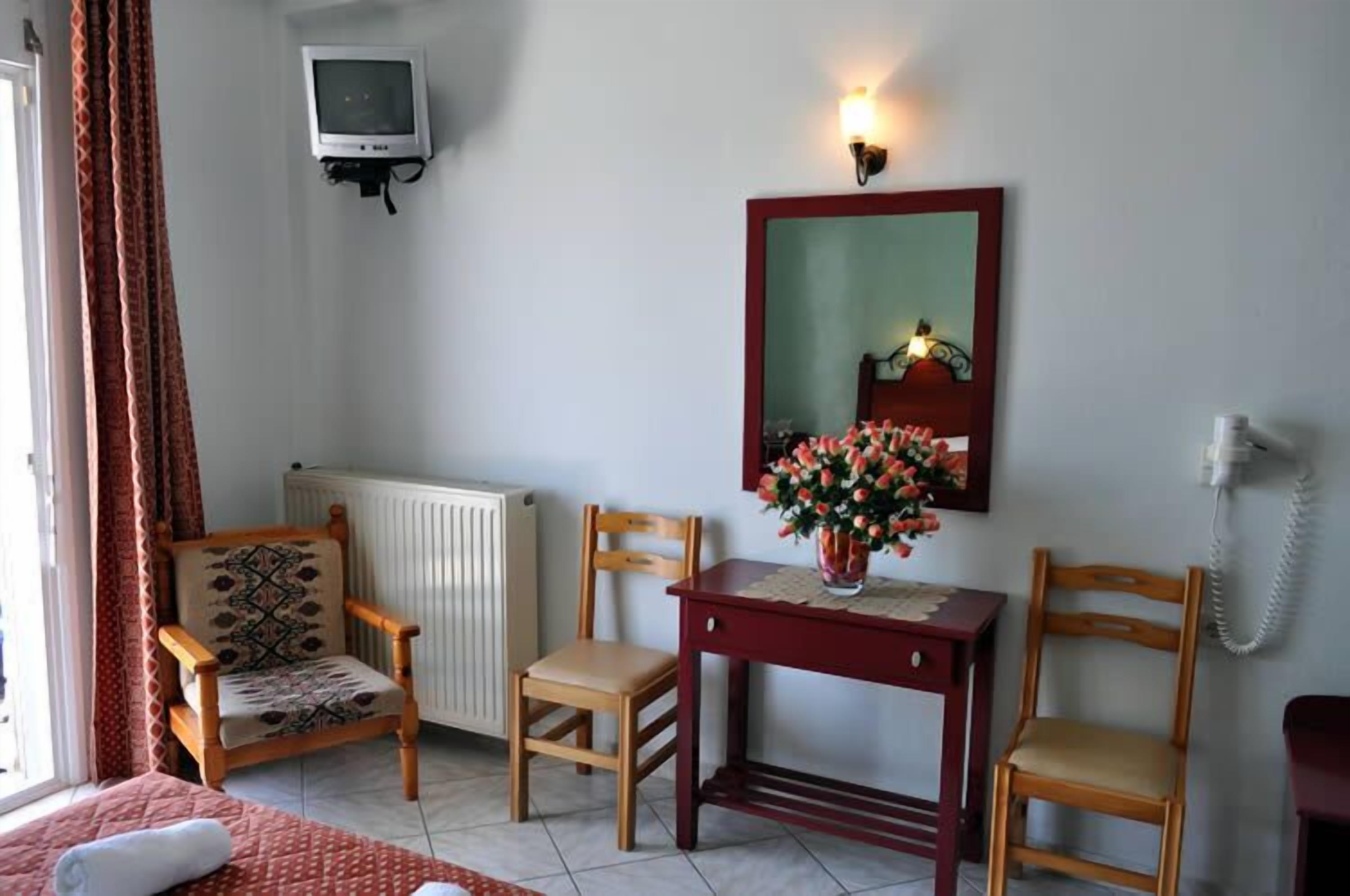 A room with a bed, two wooden chairs, a small table with a vase of flowers, a mirror, a telephone, and a radiator.