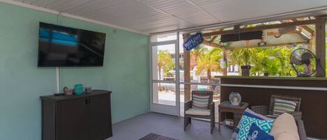 A room with a flat-screen TV, a dark-colored cabinet, a wicker sofa with patterned cushions, and a view of outdoor palm trees through a glass door.