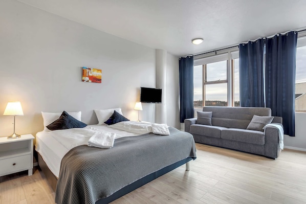 Une chambre d’hôtel avec un lit, un canapé, une télévision, un tableau et une fenêtre avec des rideaux.