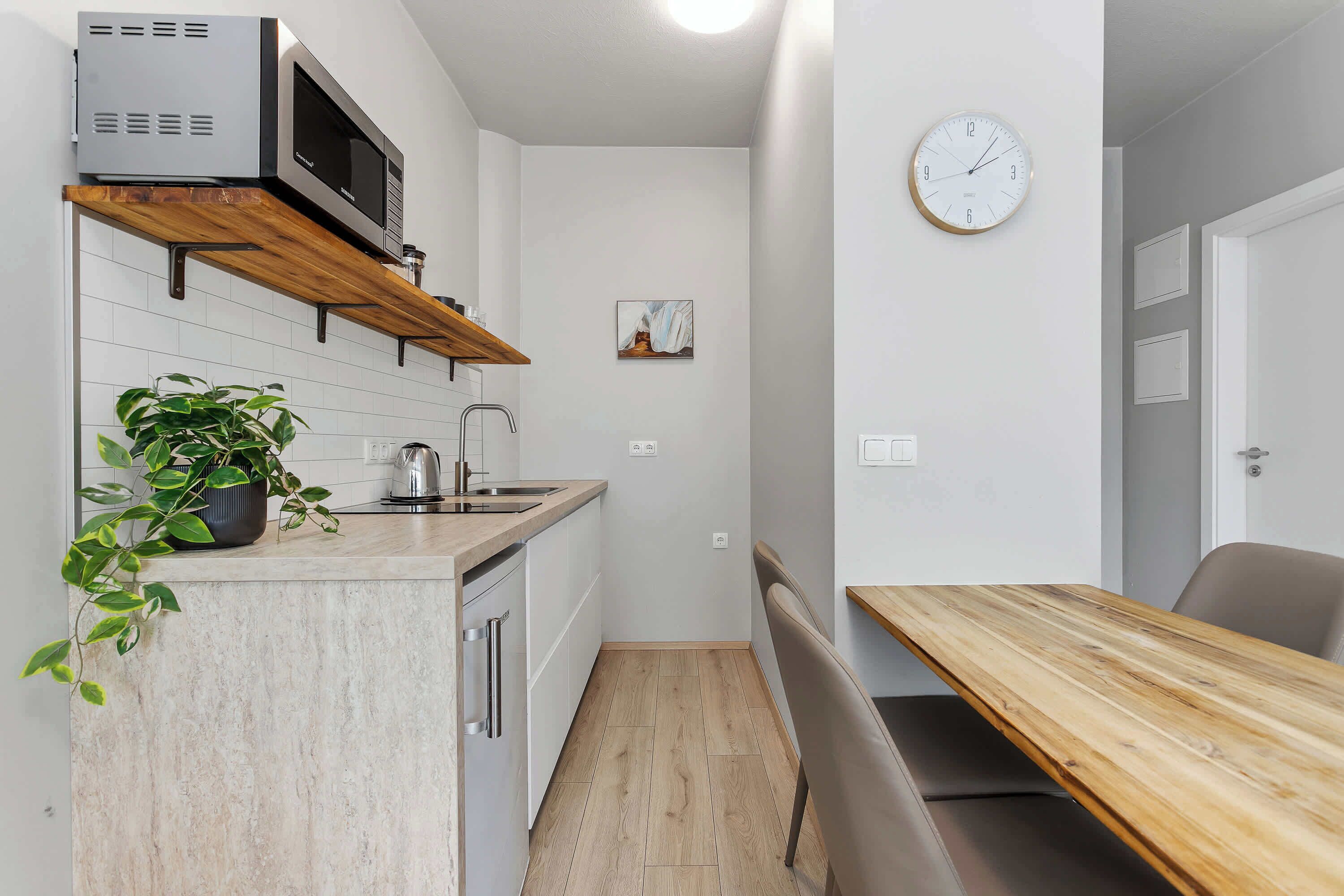 Een kleine keuken met een magnetron, een spoelbak en een houten plank. Er hangt een klok aan de muur en staat een plant op het aanrecht.