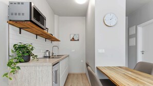 A compact kitchen with a microwave, a sink, and a wooden shelf. There is a clock on the wall and a plant on the counter.
