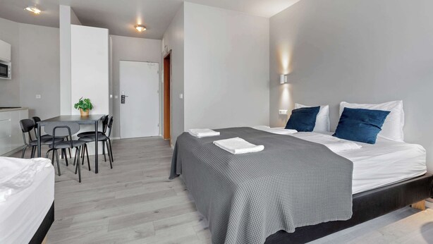 A modern hotel room with a bed, bedside tables, a desk with chairs, a potted plant, and a door.