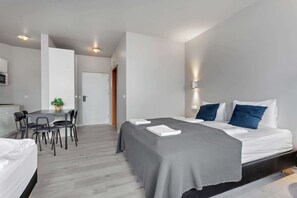 Una habitación de hotel moderna con cama, mesitas de noche, un escritorio con sillas, una planta en maceta y una puerta.