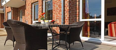 An outdoor patio with wicker chairs and a table, a brick building with large windows, and a sliding glass door.