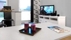 Un salón moderno con un televisor de pantalla plana, un mueble blanco para equipos electrónicos, una mesa de comedor con una bandeja de velas y vistas a un edificio.
