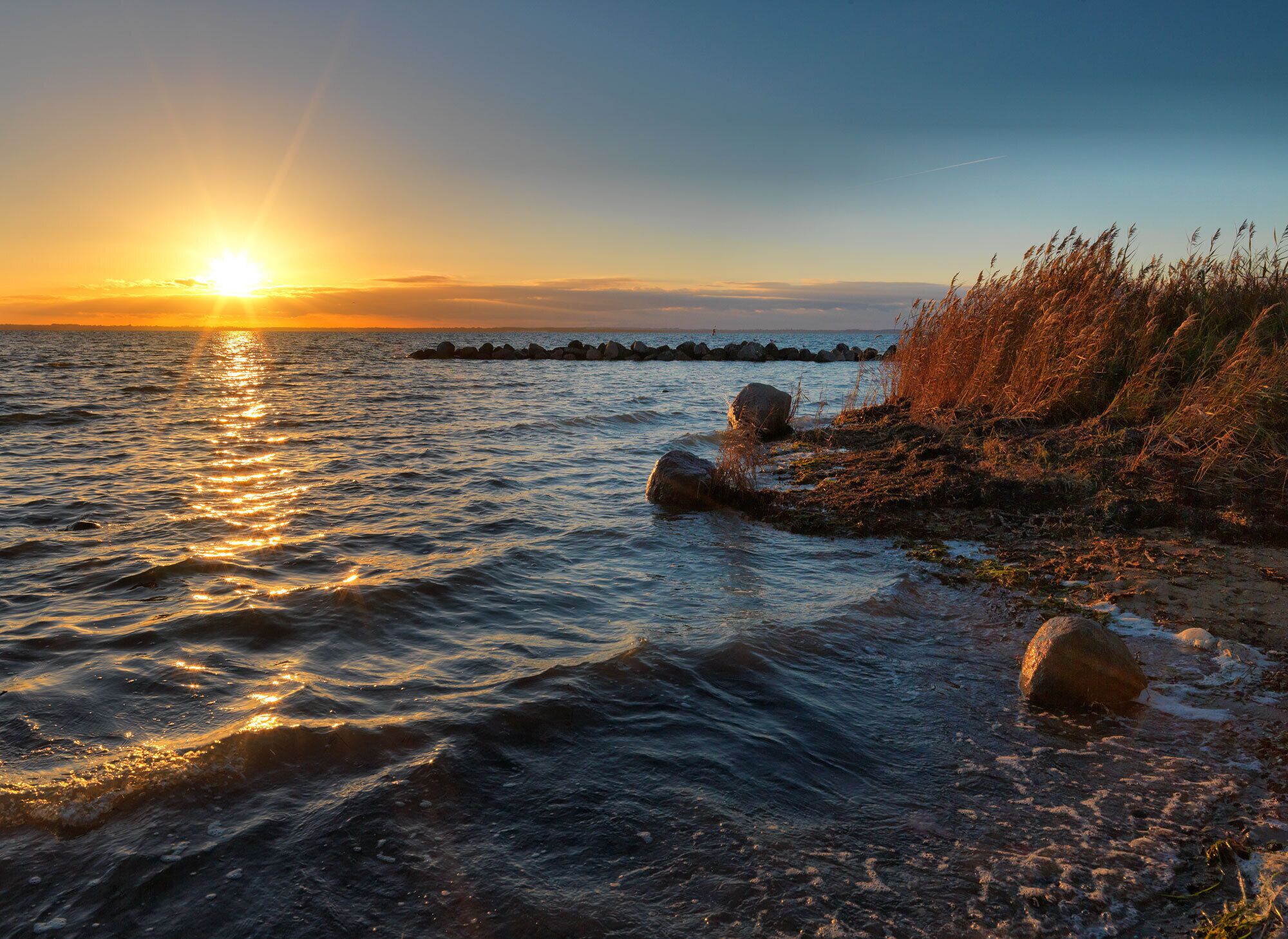 Un coucher de soleil sur une mer calme, avec des rochers et de l’herbe sur la plage.