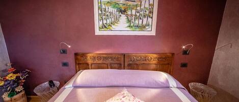 A bedroom with a wooden headboard, a bed with a floral pillow, a painting of a landscape on the wall, and two wall-mounted lamps.
