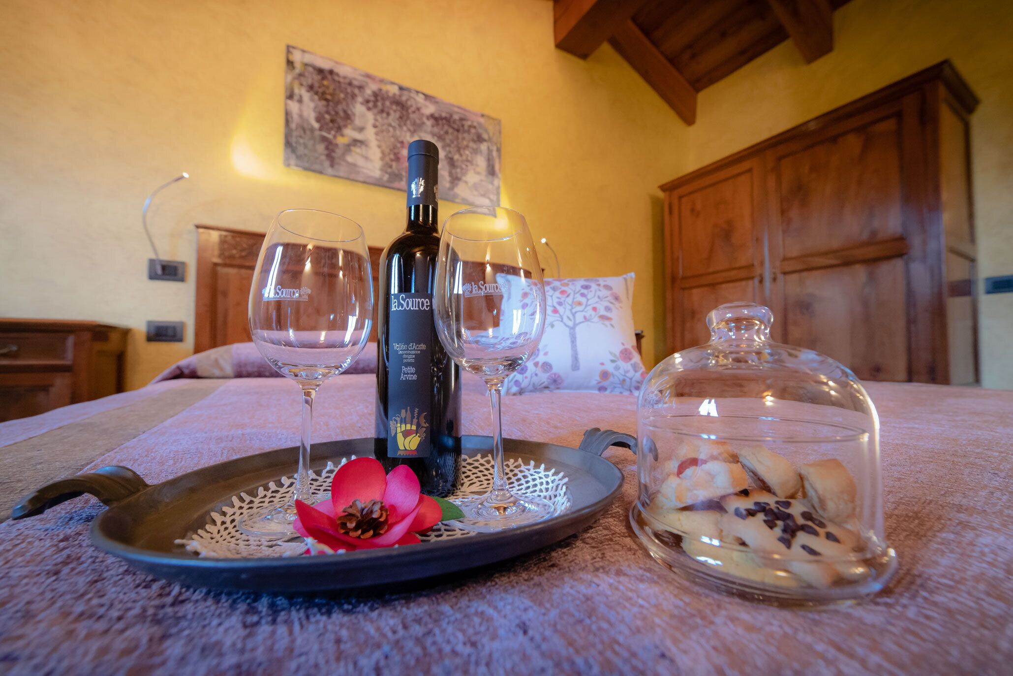 Una bandeja con una botella de vino, dos copas y un recipiente de vidrio con pasteles sobre la cama en una habitación con vigas de madera y un cuadro en la pared.