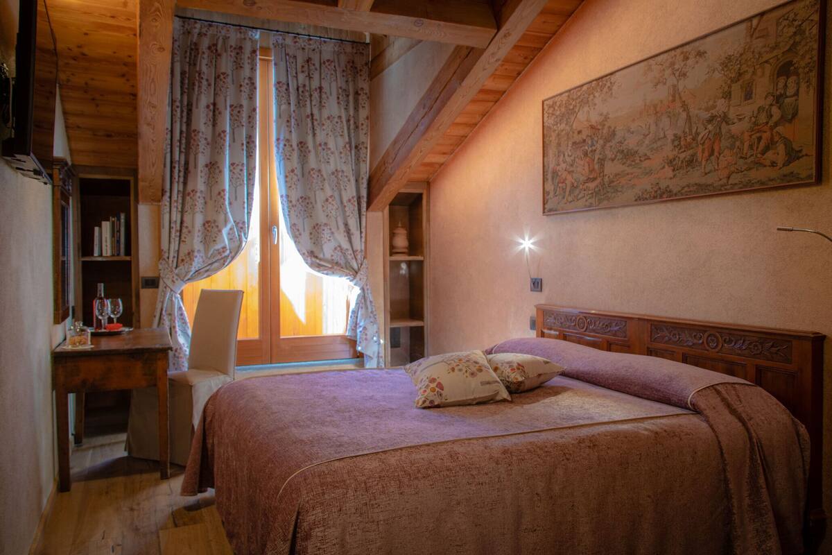 Ein Schlafzimmer mit Holzbalkendecke, einem Bett mit floralem Überwurf, einem Stuhl, einem Schreibtisch mit Büchern und Brillen sowie einem Fenster mit Vorhängen.