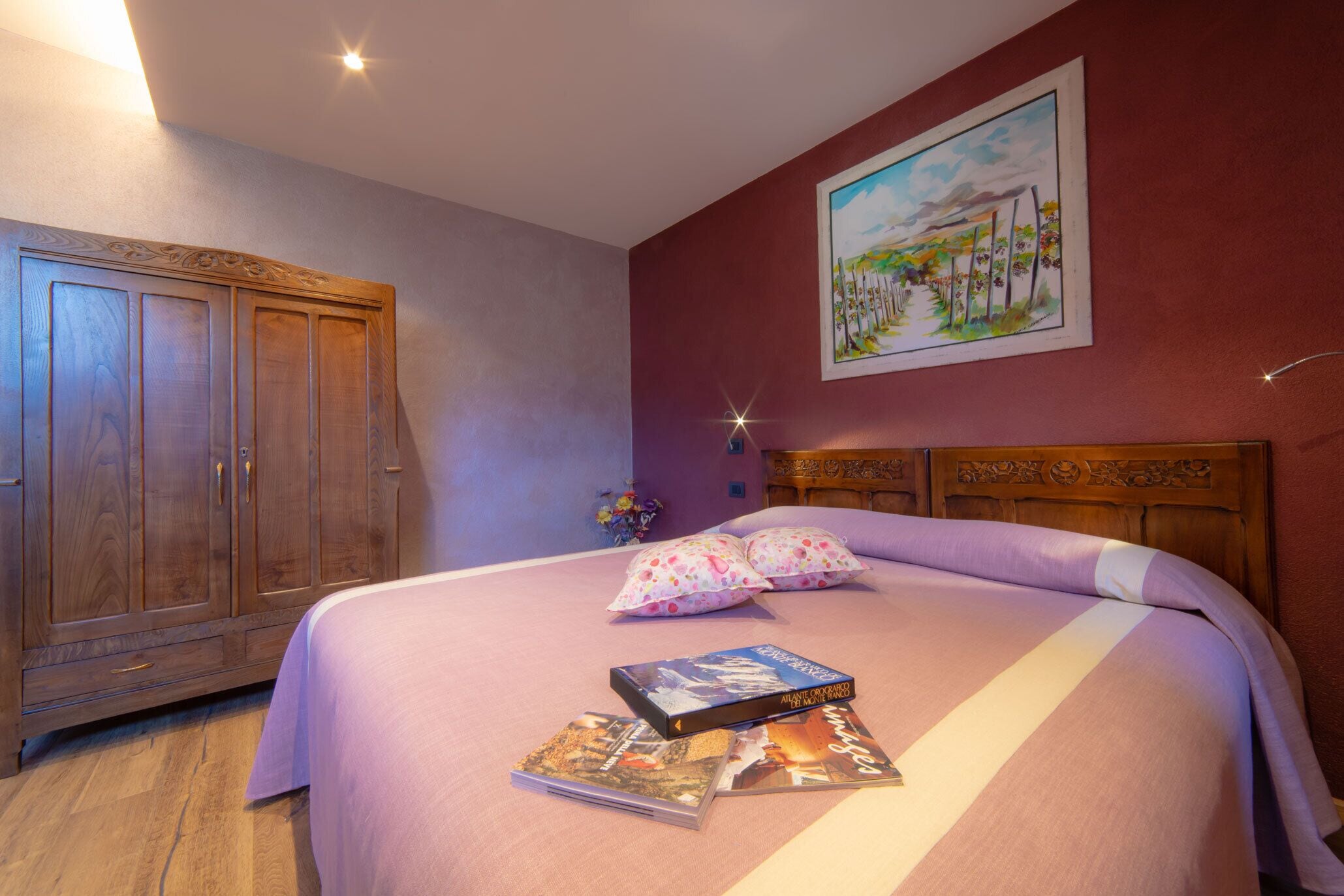 A bedroom with a wooden wardrobe, a bed with a purple bedspread, two pillows, and a painting on the wall.