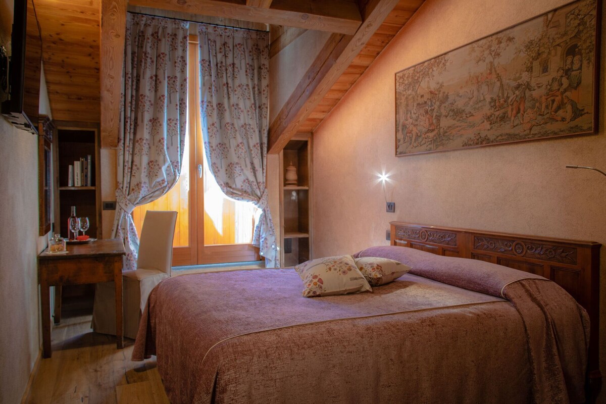 A bedroom with a wooden ceiling, a bed with a headboard, a window with curtains, a bookshelf, and a painting on the wall.