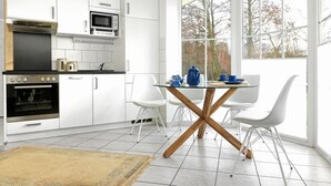 Una cocina moderna con armarios blancos, horno empotrado, microondas y comedor con mesa de vidrio y sillas blancas.