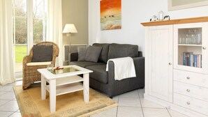 Una sala de estar con un sofá gris, una silla de mimbre, una mesa de centro blanca, una lámpara de pie y un aparador con cristalería.