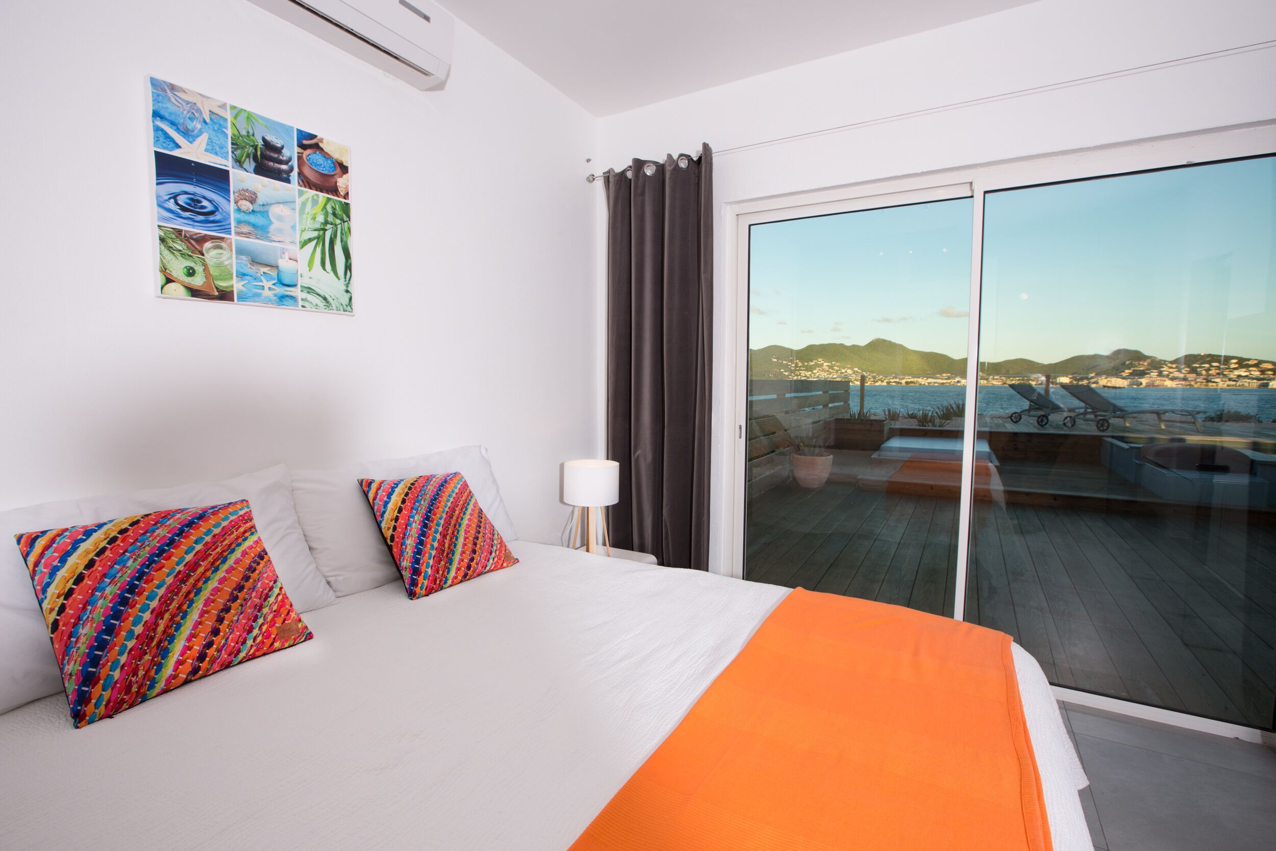 A hotel room with a bed, colorful pillows, a nightstand, a lamp, and a large window with a view of a coastal area.