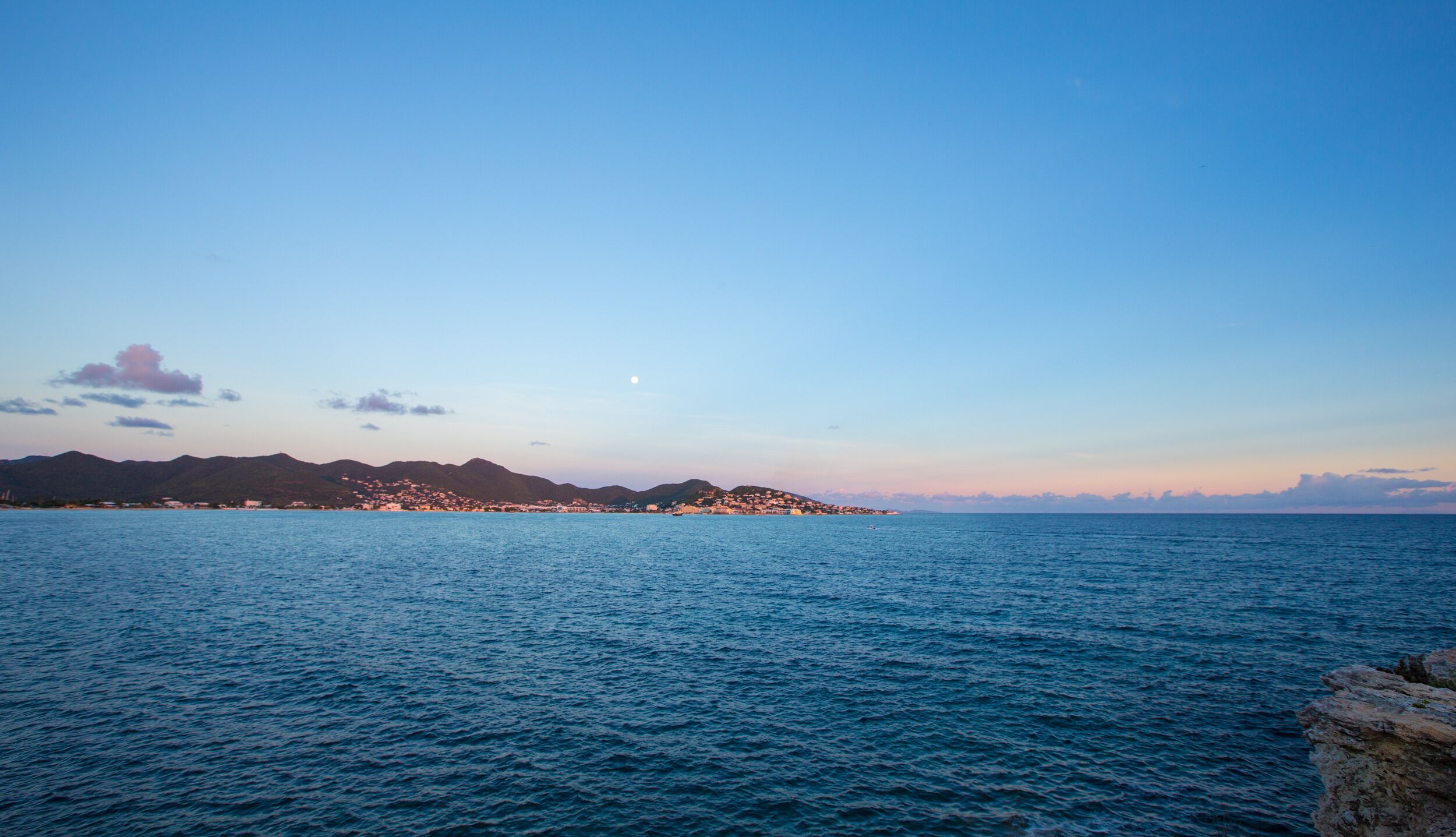 A coastal view with calm sea, distant mountains, and a clear sky.