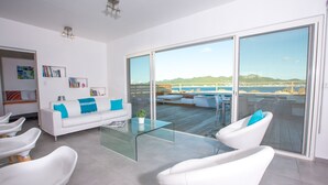 Un salón moderno con una mesa de centro de vidrio, sofás blancos con cojines azules y un amplio ventanal corredero que da a una terraza exterior con vistas al mar y las colinas.
