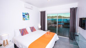 Habitación de hotel con cama, puerta de vidrio que da a un balcón con vistas al mar, televisor montado y pared decorada con un collage de fotografías.