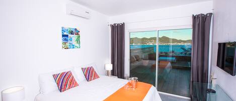 A hotel room with a bed, a glass door leading to a balcony with a view of the sea, a mounted TV, and a wall with a collage of photos.