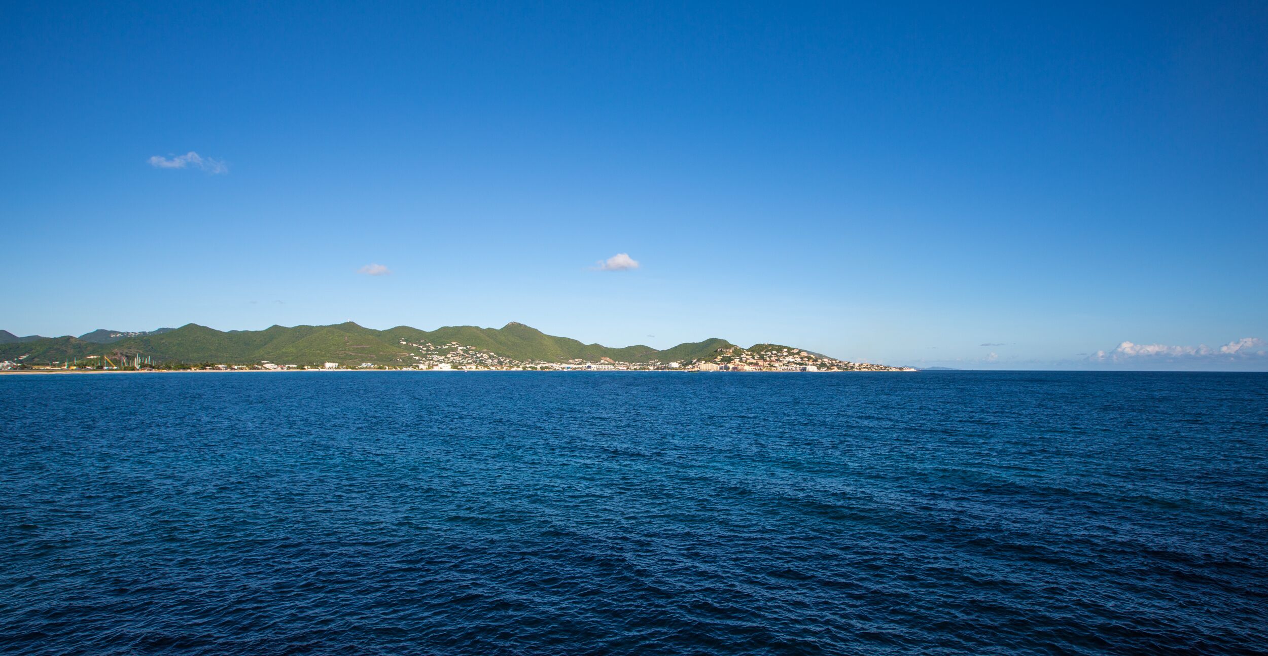 A coastal landscape with a clear sky, calm sea, and a distant hill with buildings.