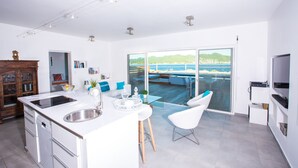 Una cocina moderna con una isla grande, un comedor con mesa de vidrio y vistas al mar a través de una puerta corrediza de cristal.