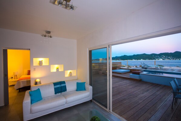 Un moderno salón con un sofá blanco, una mesa de centro de vidrio y vistas a una terraza de madera con mobiliario exterior.