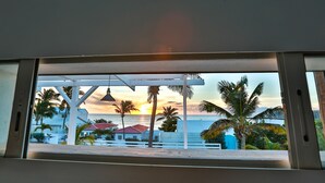 Vista a través de una ventana de un edificio con barandal blanco, palmeras y un atardecer sobre el océano.